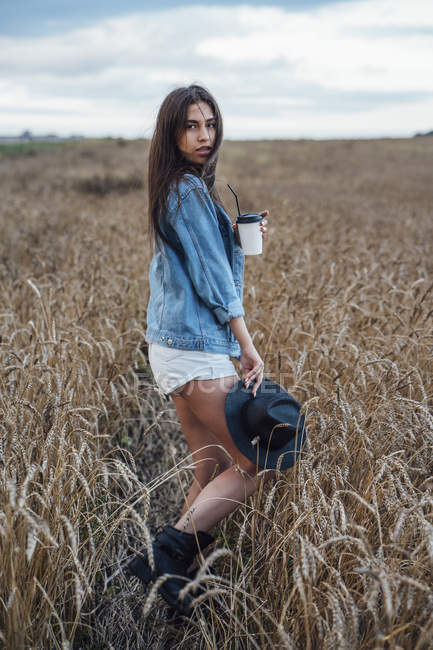 Portrait of young woman with beverage walking in corn field — solitude ...