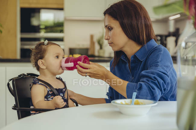 Mother helping baby daughter drinking water from bottle in kitchen — Stock Photo