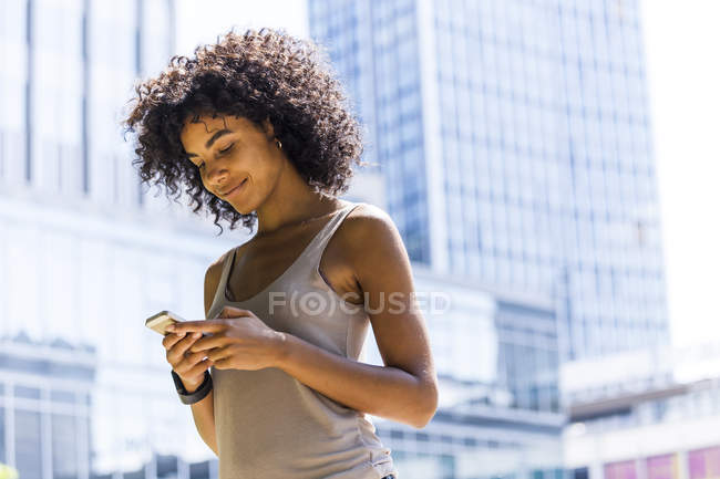 Jovem sorridente com cabelo encaracolado usando telefone celular na frente de arranha-céus — Fotografia de Stock