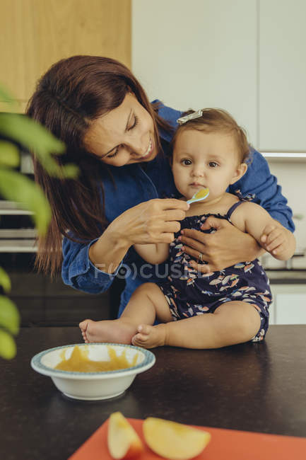 Mother helping baby daughter eating fruit pulp in kitchen — Stock Photo
