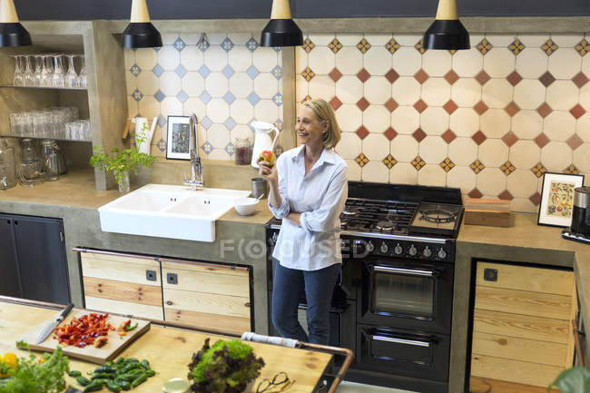 Улыбающаяся взрослая женщина держит яблоко на кухне — стоковое фото