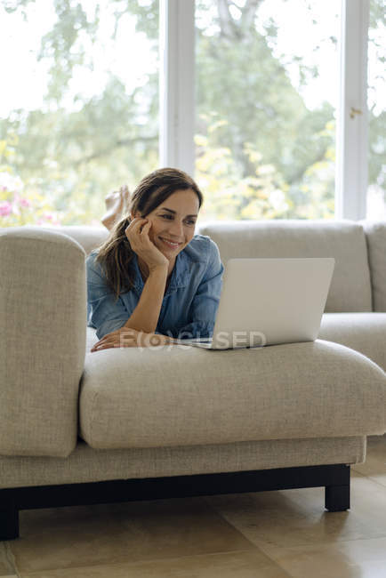 Sorrindo mulher madura deitada no sofá em casa usando laptop — Fotografia de Stock