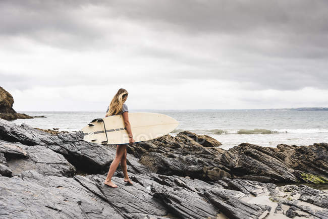 Giovane donna che porta la tavola da surf sulla spiaggia rocciosa in mare — Foto stock