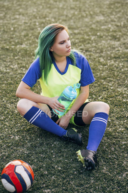 Mujer joven sentada en el campo de fútbol con botella de agua y pelota - foto de stock