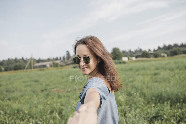 Femme souriante portant des lunettes de soleil tenant la main d'un partenaire dans un champ rural — Photo de stock