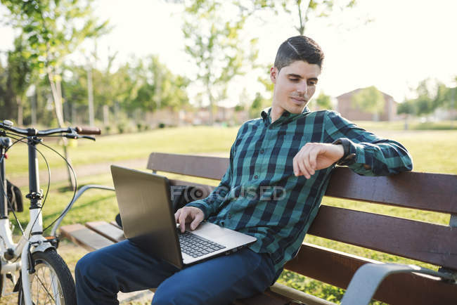 Jeune homme avec ordinateur portable sur banc de parc temps de contrôle — Photo de stock