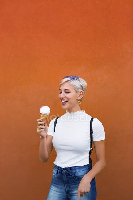 Mujer joven riendo con cono de helado en frente de fondo naranja - foto de stock