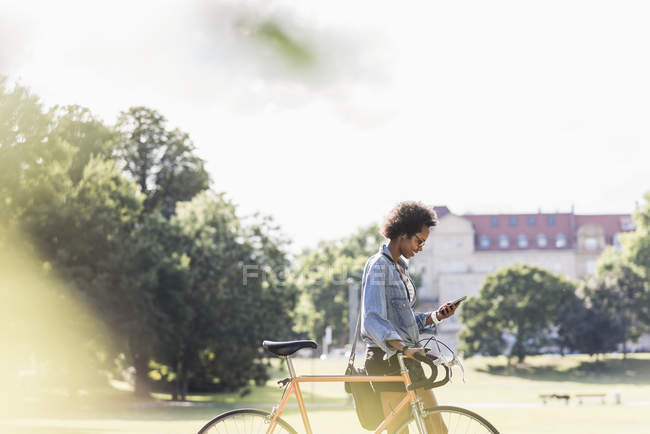 Giovane donna con cellulare spingendo bicicletta nel parco — Foto stock