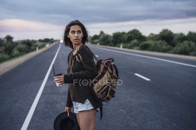 Retrato de una joven autoestopista con mochila y bebida de pie en el carril - foto de stock