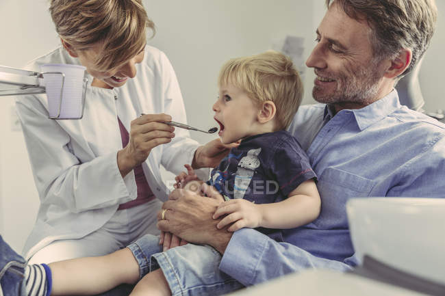Жіночий стоматолог оглядає маленького хлопчика, сидячи на колінах батька. — стокове фото