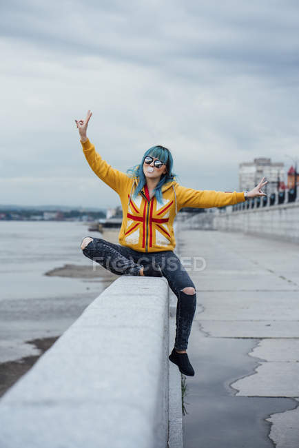 Retrato de mulher jovem com cabelo azul tingido sentado na parede mostrando sinais de vitória — Fotografia de Stock