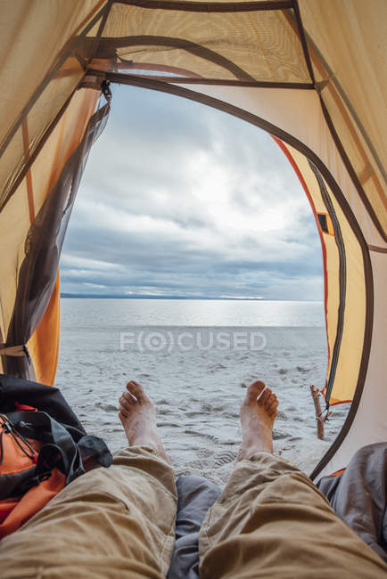 Uomo scalzo sdraiato in tenda sulla spiaggia al lago acqua e cielo nuvoloso — Foto stock