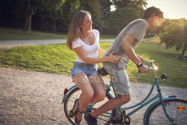 Jeune couple à vélo dans le parc, femme assise sur un rack — Photo de stock