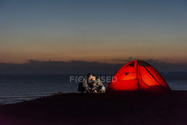 Романтична пара кемпінг на пляжі, використовуючи смартфон у сутінках — стокове фото