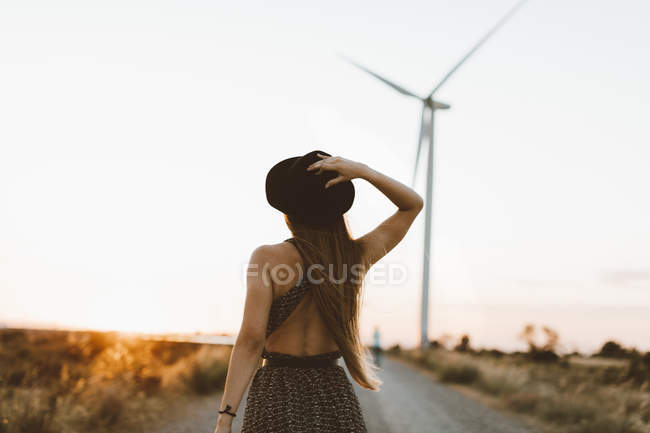 Rückansicht einer jungen Frau auf einer Landstraße am Abend mit einem Windrad im Hintergrund — Stockfoto