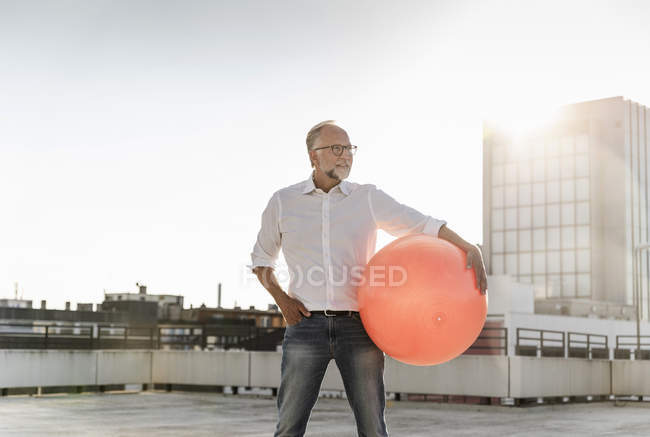 Älterer Mann spielt mit orangefarbenem Fitnessball auf dem Dach eines Hochhauses — Stockfoto