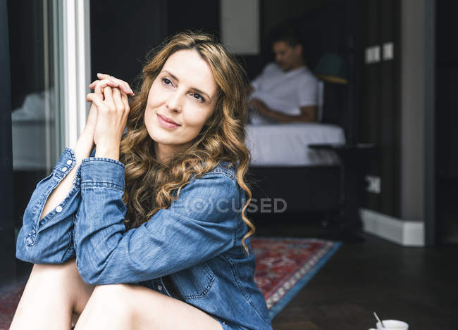 Femme souriante assise à la porte-fenêtre avec l'homme en arrière-plan — Photo de stock