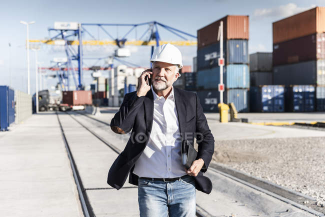Бизнесмен в грузовой гавани, в защитном шлеме, со смартфоном — стоковое фото