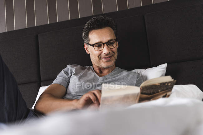 Smiling man lying in bed at home reading a book — Stock Photo