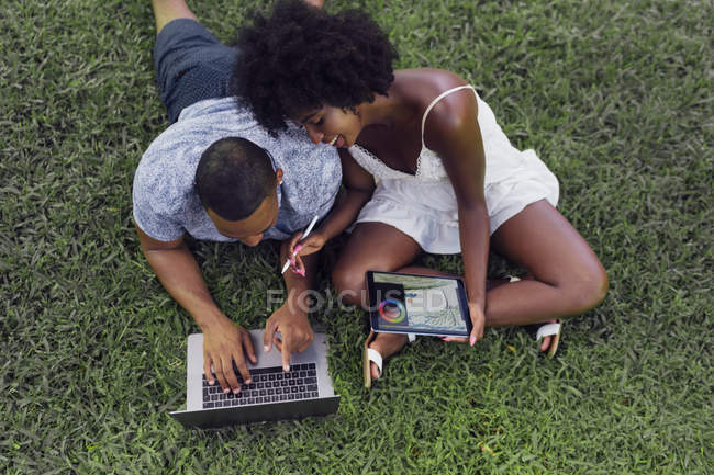 Jeune couple utilisant tablette et ordinateur portable sur la pelouse dans un parc — Photo de stock