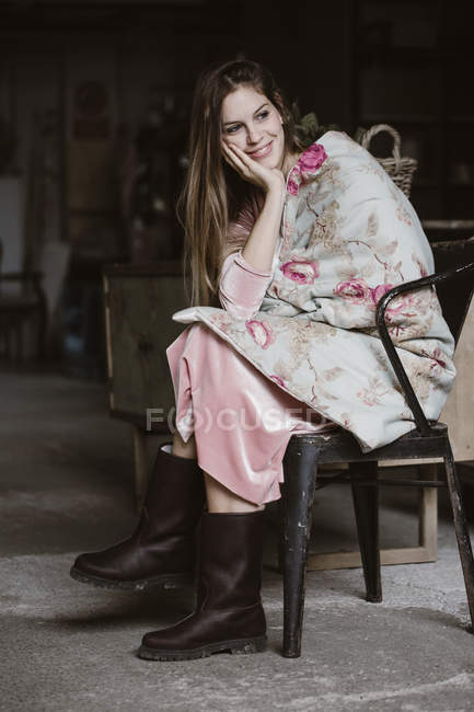 Портрет усміхненої молодої жінки, що сидить на горищі, загорнутої в ковдру з квітковим дизайном — стокове фото
