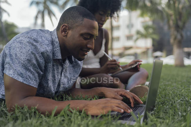USA, florida, miami beach, junges paar mit tablet und laptop auf rasen in einem park — Stockfoto