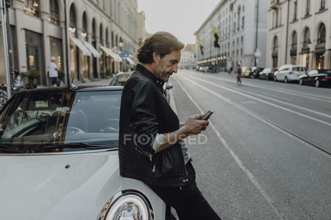 Reifer Mann lehnt am Auto, hält Smartphone, München, Bayern, Deutschland — Stockfoto