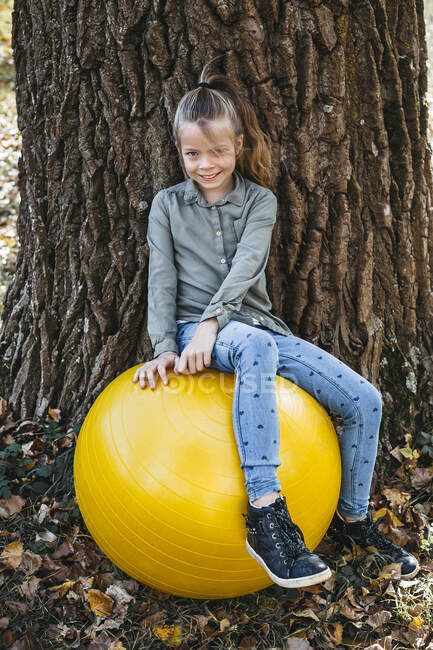 Ritratto di ragazza sorridente seduta sulla palla gialla della palestra appoggiata al tronco d'albero — Foto stock
