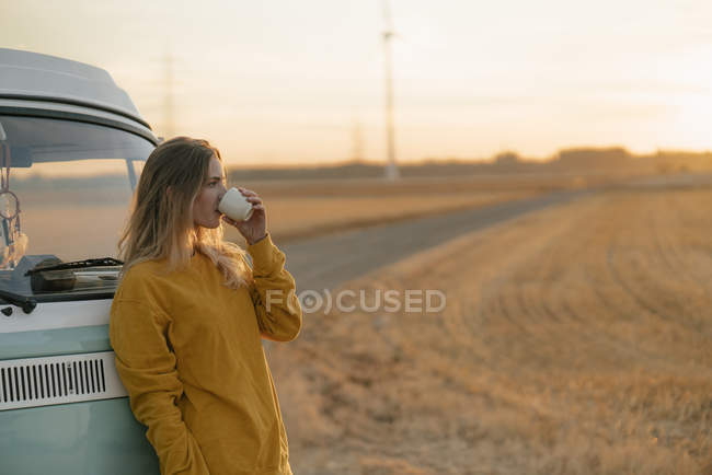 Молода жінка п'є з кухля в кемпер Ван в сільській місцевості на заході сонця — стокове фото