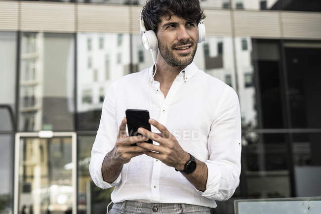 Молодий чоловік з навушниками та мобільним телефоном у місті, дивлячись далеко — стокове фото