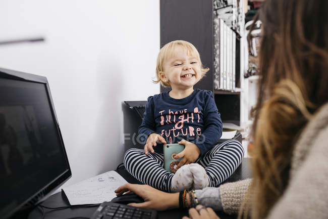 Madre trabajando en casa con su hijita sentada en el escritorio - foto de stock