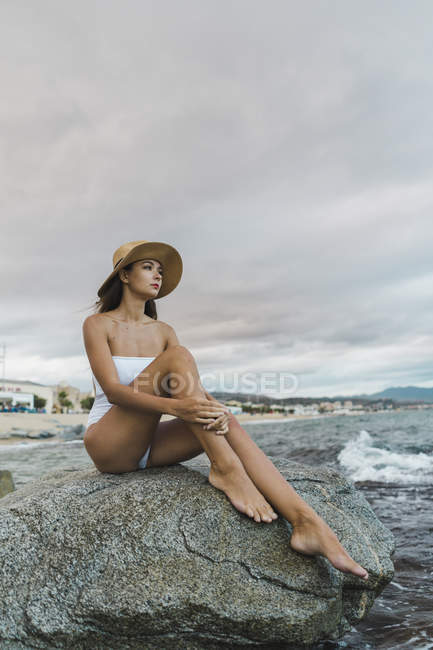 Bella giovane donna che indossa costume da bagno e cappello, seduta sulla roccia nel mare — Foto stock