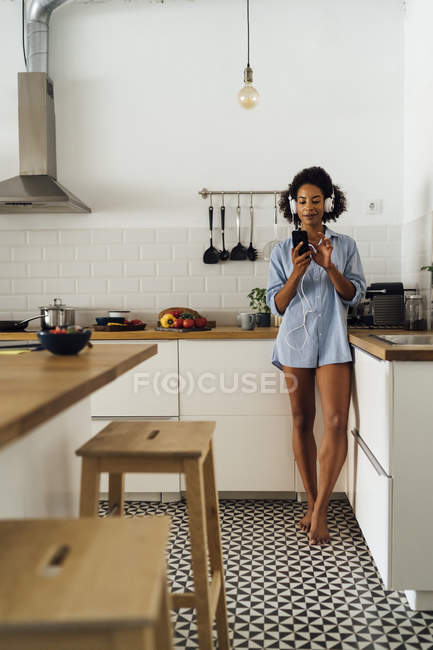 Жінка з навушниками, використовуючи смартфон в її кухні — стокове фото