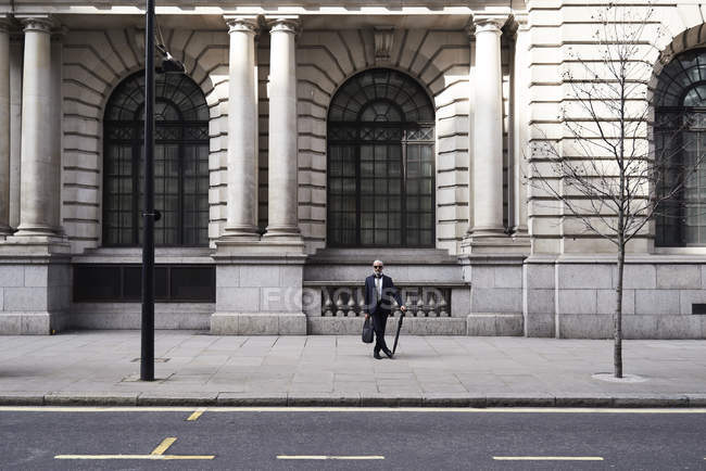 Великобритания, Лондон, стильный высокопоставленный бизнесмен с чемоданом и пупсом, стоящий на тротуаре — стоковое фото