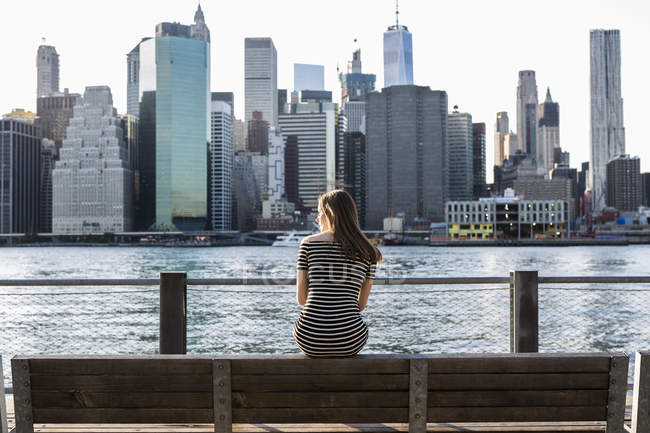 Estados Unidos, Nueva York, Brooklyn, vista trasera de la mujer sentada en el banco frente a East River y el horizonte de Manhattan - foto de stock