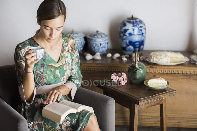 Young woman drinking cup of tea while reading a book at home — Stock Photo