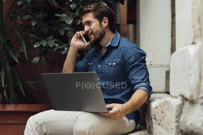 Усміхнений бізнесмен по телефону сидить на сходах у дворі з ноутбуком — стокове фото