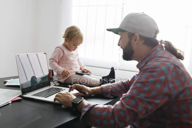 Vater arbeitet von zu Hause aus, benutzt Laptop, während seine Tochter auf dem Schreibtisch sitzt und spielt — Stockfoto