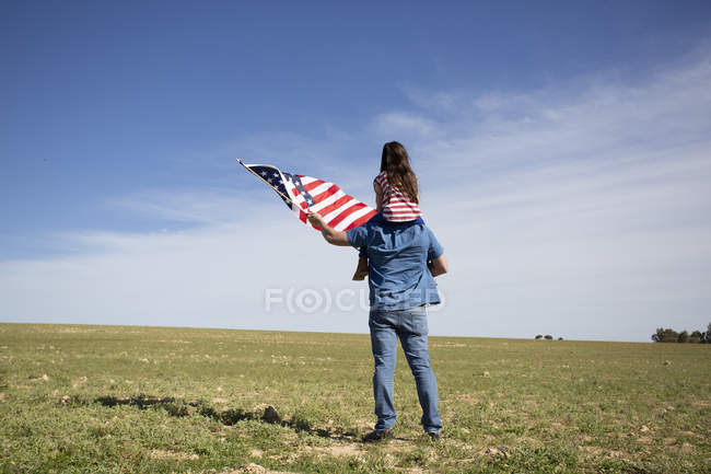 Un homme avec sa fille et un drapeau américain debout dans un paysage éloigné — Photo de stock