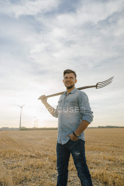 Ritratto di giovane uomo sorridente che tiene il forcone in piedi sul campo di stoppie — Foto stock