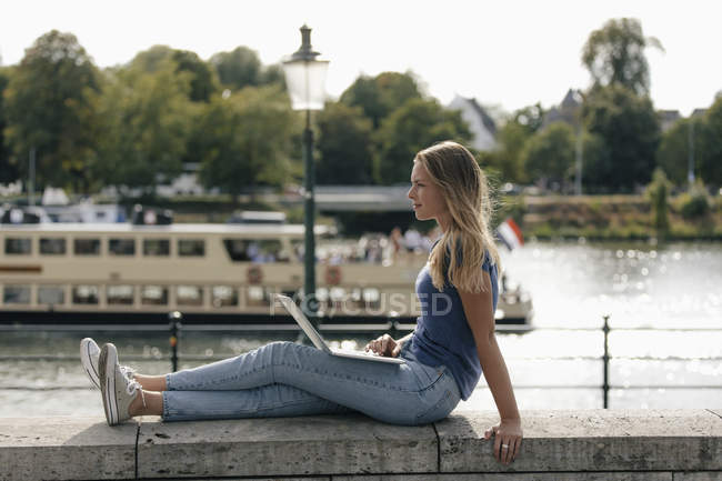 Нідерланди, Маастрихт, молода жінка, сидячи на стіні на березі річки з ноутбуком — стокове фото