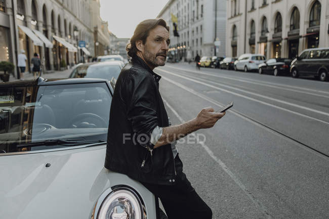 Зрілий чоловік, який спирається на машину, тримає смартфон (Мюнхен, Баварія, Німеччина). — стокове фото