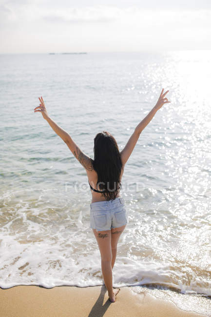 Вид ззаду молода жінка стоїть на березі моря і показує знак перемоги — стокове фото