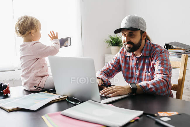 Padre che lavora da casa, usando il computer portatile con la figlia seduta sulla scrivania, giocando — Foto stock