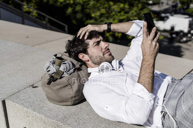 Jeune homme couché sur le mur à l'extérieur et utilisant un téléphone portable — Photo de stock
