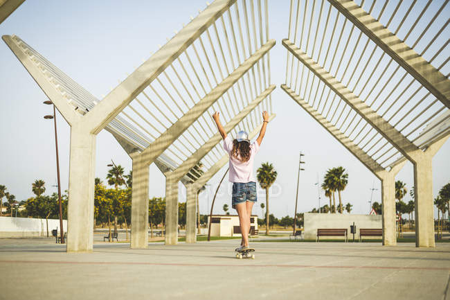 Девушка на скейтборде, с поднятыми руками, вид сзади — стоковое фото