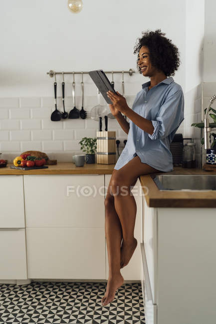 Frau sitzt auf Arbeitsplatte ihrer Küche und benutzt morgens ein digitales Tablet — Stockfoto