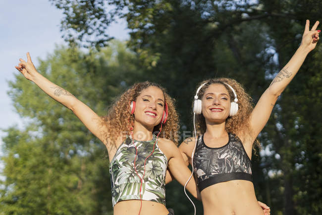 Retrato de hermanas gemelas sonrientes escuchando música con auriculares y mostrando signo de victoria - foto de stock