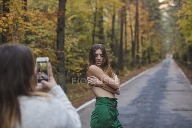 Woman taking cell phone picture of barechested young woman standing on country road — Stock Photo
