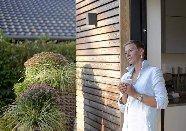 Donna pensierosa con una tazza di caffè in piedi davanti alla sua casa — Foto stock